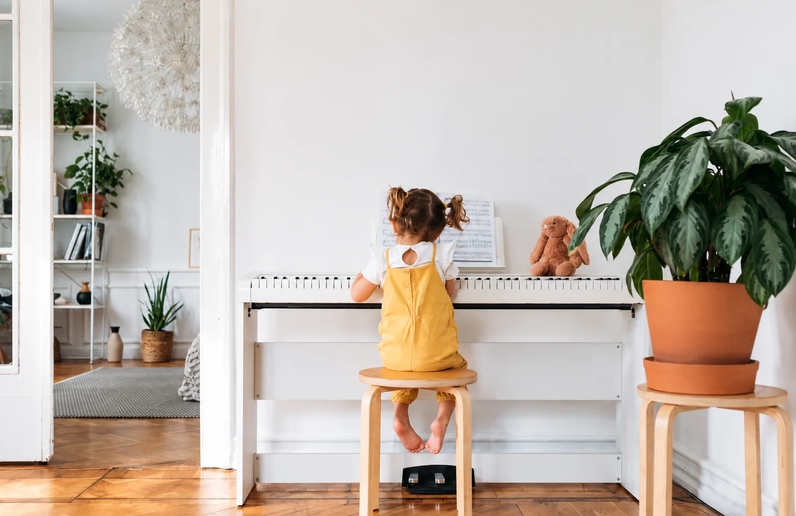 A young child seated at a white piano beside a houseplant